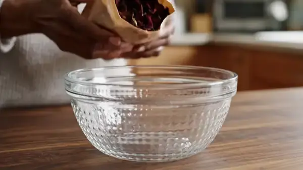 Weathered hands pouring dried red hibiscus calyces from a plain paper bag into a glass bowl